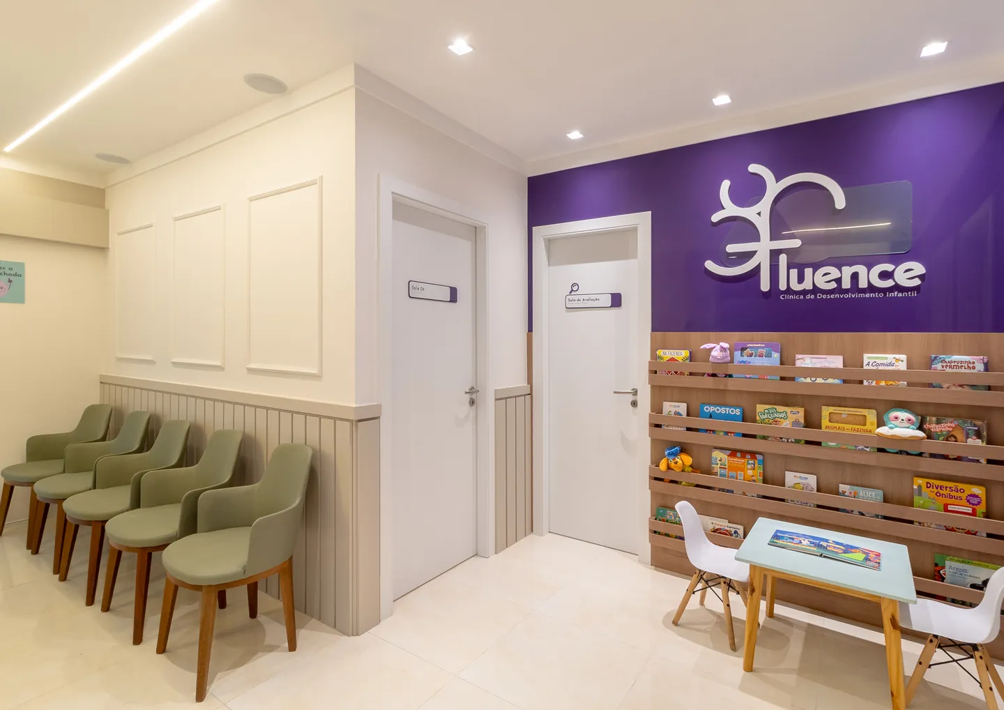 Espera da Clínica Fluence: poltronas verdes, boiserie branca, parede roxa com o letreiro da clínica e estante de livros infantis com mesinha.